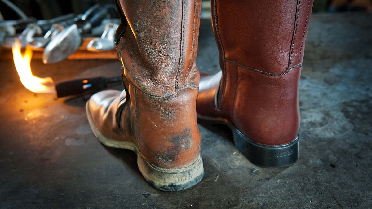 A close-up of two Petrie riding brown leather boots from behind, a dirty one and a clean one, in a workshop setting with a forge flame visible in the background.