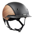 A matte black equestrian riding Casco helmet with natural wood-look panels on the sides, featuring a black chin strap and a visible 'CASCO' brand, 3/4 front view against white background