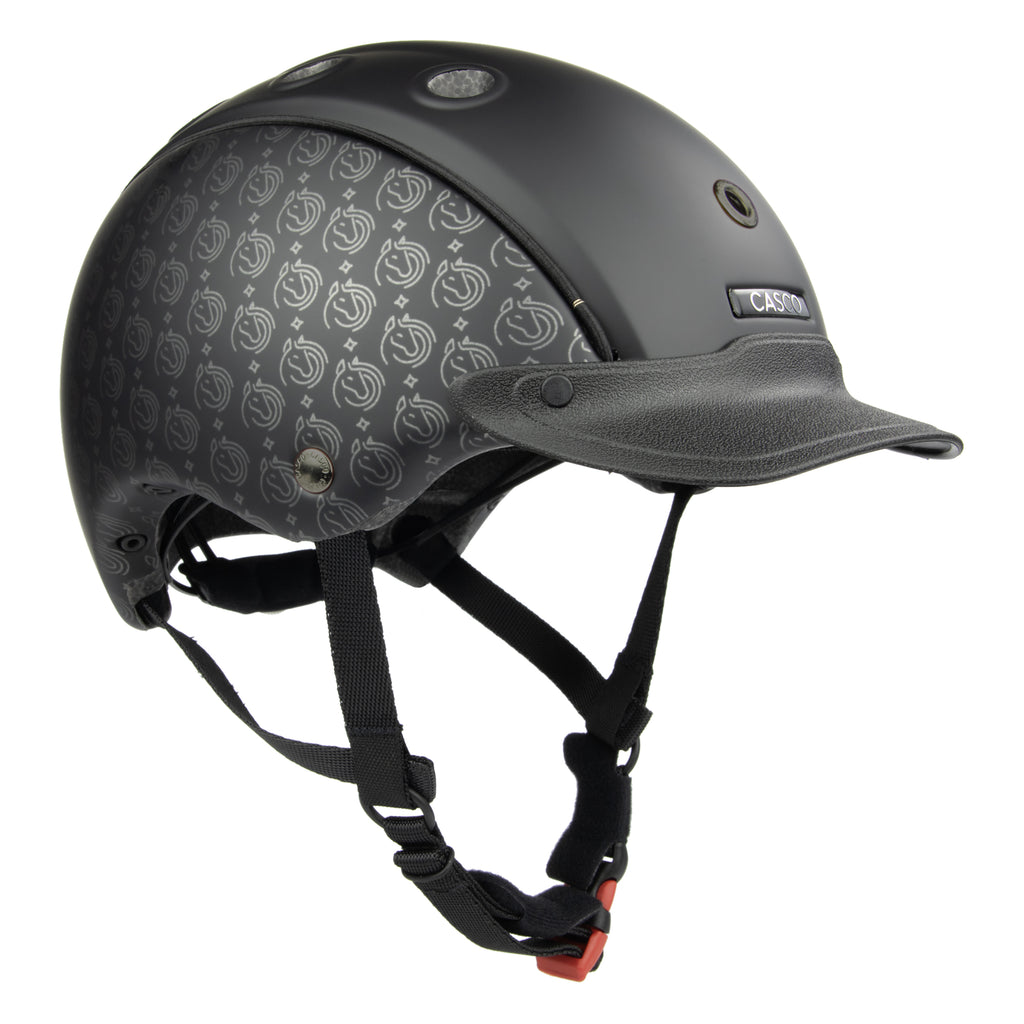 A CASCO matte black equestrian helmet with a subtle repeating logo pattern on the side panels, a matching black visor, and black chin straps with an orange release buckle, shown on a solid white background.
