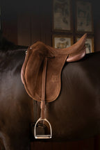 A brown leather Childeric DPL dressage saddle with a matching leather girth and silver metal stirrups, shown mounted on the back of a dark brown horse in a dimly lit indoor equestrian setting.