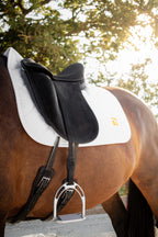 A close-up side view of a brown horse wearing a Childeric dressage black saddle with a Childeric white saddle pad and stirrups, outdoors with trees and bright sunlight in the background.