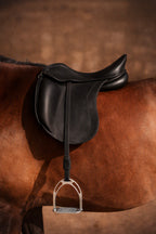A close-up of a black leather Childeric dressage saddle with a silver metal stirrup, attached to a brown horse against a warm brown background.