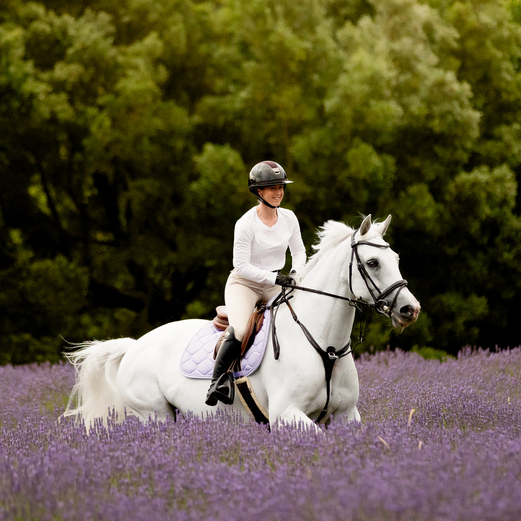 A woman  riding a white horse with a Lumiere Milan convertible snaffle bridle through a lavender field, with a dense green forest in the background.