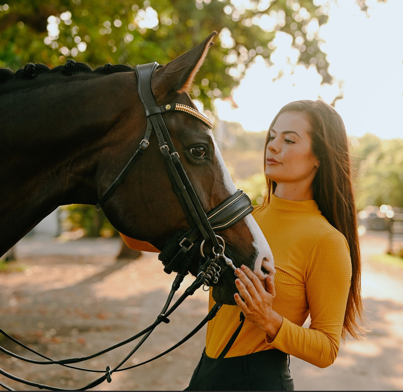 Woman in a yellow sweater with a Brown horse wearing the Lumiere Delta double bridle crafted from premium leather with gold browband features a clincher-style centrepiece encased by gold beading, the noseband is gel-padded and finished with gold piping 
The gel-padded anatomic poll-relief headpiece - a blurred green background