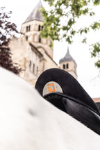 A close-up of a Childeric DPL Dressage black leather saddle with the Childeric logo, on a white horse's back, with a blurred stone church tower and green tree branches in the background.