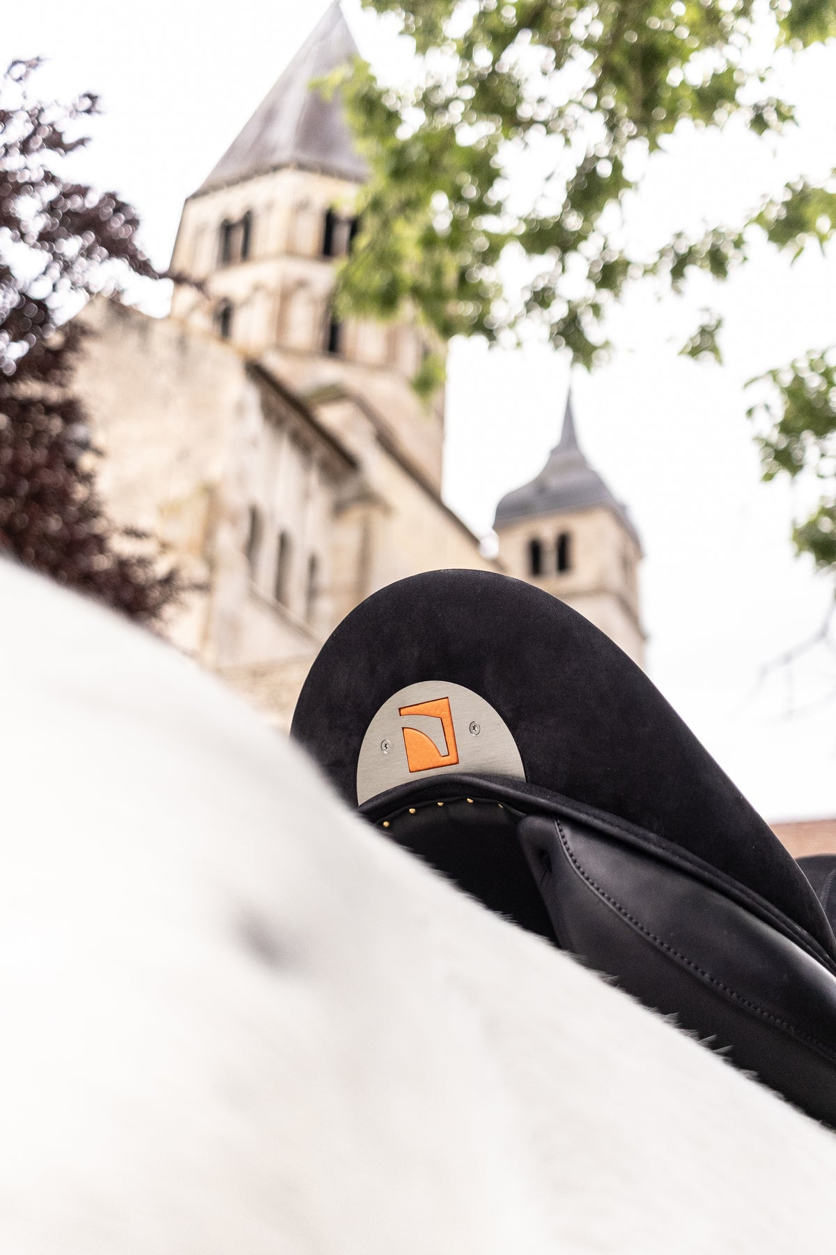 A close-up of a Childeric DPL Dressage black leather saddle with the Childeric logo, on a white horse's back, with a blurred stone church tower and green tree branches in the background.