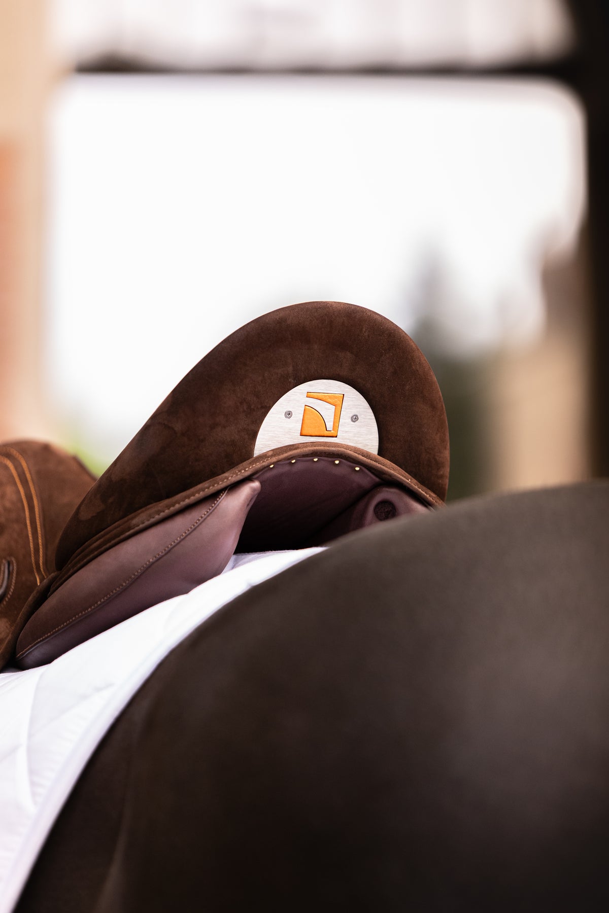 A close-up shot of a brown Childeric DPL dressage saddle  with a white and orange Childeric logo patch, on a white saddle pad and dark horse back.