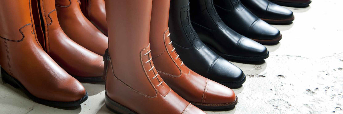 Several pairs of all-round Petrie riding boots in black and Havana colours align on a concrete ground